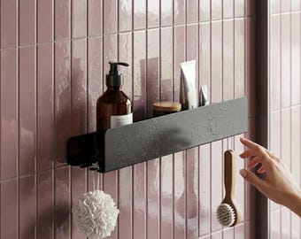 May include: A matte black rectangular bathroom shelf mounted on a mauve tiled wall. The shelf holds a brown pump bottle, a small jar, and two tubes. A white bath sponge and a wooden handled brush hang from the bottom edge of the shelf.