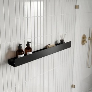 Könnte beinhalten: Ein schwarzes Metallregal an einer weißen gefliesten Wand, das zwei braune Flaschen mit weißen Etiketten, eine kleine Bürste und eine schwarze Vase mit Schilf enthält. In einem Badezimmer.