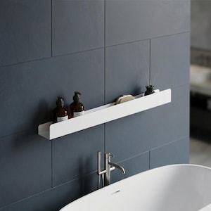 May include: A long, narrow, white metal wall-mounted shelf featuring a flat base and a tall front edge. The shelf holds two amber-colored glass bottles with black pump dispensers, a small beige oval brush, and a black reed diffuser bottle.