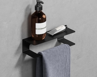 May include: A matte black metal wall-mounted shelf featuring a flat top surface for storage and a lower bar for hanging towels. The shelf holds a brown amber glass pump bottle with a white label and a small silver metal tube of personal care product.