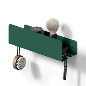 May include: A dark green wall-mounted metal shelf featuring a flat top surface holding a small black jar and a shaving brush with light-colored bristles. The shelf includes a side hook holding a round wooden brush and a slot for a black razor.