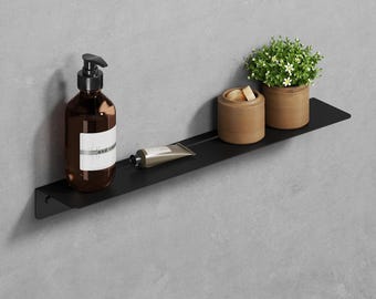 Puede incluir: Estante de metal negro con un dispensador de jabón marrón, un tubo de crema, un pequeño recipiente de madera y una planta en maceta con hojas verdes y flores blancas. El estante está montado en una pared gris.