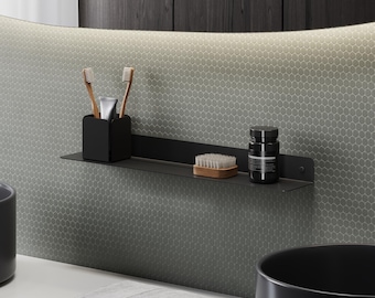 Puede incluir: Un estante de pared negro mate que sostiene un soporte para cepillos de dientes negro con dos cepillos de madera, un cepillo pequeño de madera y un recipiente cilíndrico negro con etiqueta blanca, montado sobre azulejos hexagonales grises.