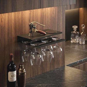 May include: A black metal wine glass rack with two shelves, holding six clear wine glasses. A gold and wood corkscrew rests on the top shelf, along with two coasters. A bottle of red wine and a cocktail shaker are in the foreground.