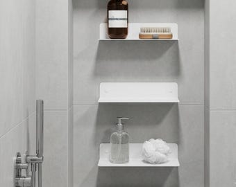 May include: Three white metal wall-mounted shelves installed in a shower niche. The top shelf holds a brown bottle and a wooden brush. The middle shelf is empty. The bottom shelf holds a clear soap dispenser and a white mesh bath sponge.