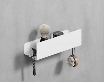 May include: A white metal wall-mounted organizer with a shaving brush, razor, small jar, and a wooden-handled brush. The organizer is designed to hold grooming essentials and has a minimalist aesthetic.