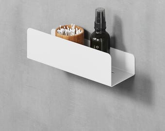 May include: A white wall-mounted shelf with a rectangular base and a front panel. On the shelf sits a small brown wooden container filled with white cotton swabs and a black spray bottle with a pump dispenser and white text on the label.