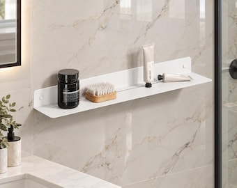 May include: A long, white, wall-mounted metal shelf featuring a thin horizontal slot. The shelf holds a black cylindrical jar, a small wooden-handled brush with light-coloured bristles, and two white tubes of skincare products.