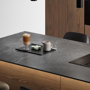 May include: A clear glass mug filled with a layered coffee beverage topped with foam, a small light green macaron, and a matte off-white ceramic creamer pitcher arranged on a rectangular black tray set upon a dark grey stone countertop.