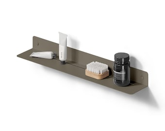 May include: A long, taupe-colored metal wall shelf with a thin horizontal slot running along its center. The shelf holds two white tubes, a small wooden brush with white bristles, and a dark brown plastic jar with a black screw-top lid.