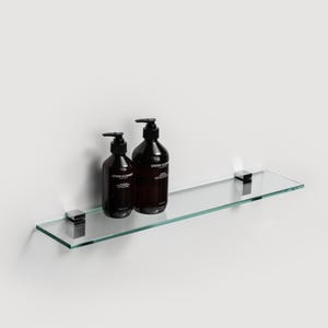 May include: A glass shelf with two brown glass bottles of hand soap with black pump tops. The shelf is mounted to a white wall with two chrome brackets.
