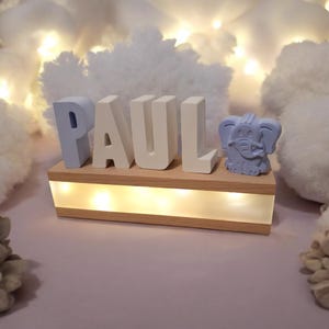 Könnte beinhalten: Ein Holz-Namensschild mit dem Namen "PAUL" in weißen und hellblauen Buchstaben. Das Schild hat eine hellblaue Elefantenfigur auf der rechten Seite und eine hellbraune Basis mit eingebauten LED-Lichtern.