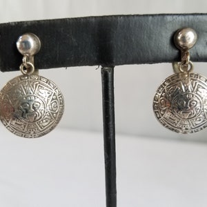 May include: A pair of silver clip-on earrings with a circular design featuring a stylized face and intricate details. The earrings are displayed on a black stand.