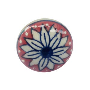 May include: A round, ceramic cabinet knob with a floral design. The knob features a white flower with blue outlines and a dark blue center, set against a red background with white polka dots. The knob is a decorative element for furniture.