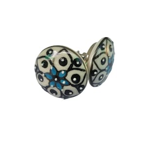 May include: Two round earrings with a floral design. The earrings have a cream-colored background with black and blue floral patterns. The earrings are made of a shiny material and have a silver-colored backing.