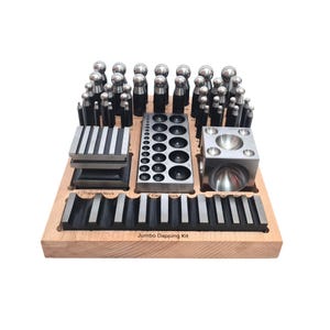 May include: A wooden jewelry-making kit with various metal tools. The kit includes dapping blocks, a forming block, and a variety of metal punches and forming tools. The text "Jumbo Dapping Kit" is printed on the wooden base.