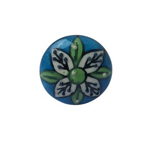 May include: A round, blue ceramic knob with a white and green floral design. The flower has white petals with black detailing, green leaves, and a green center. The knob is likely for a cabinet or drawer.
