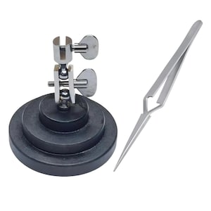 May include: A watch holder with a black base and a silver metal clamp, along with a pair of silver tweezers. The watch holder has adjustable clamps with wingnuts. The tweezers have pointed tips.