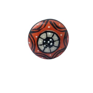 May include: A round, decorative ceramic knob with a vibrant orange background. The knob features a central design of white and black concentric circles, surrounded by black curved lines and small white dots. This unique knob is perfect for adding a pop of color to furniture.