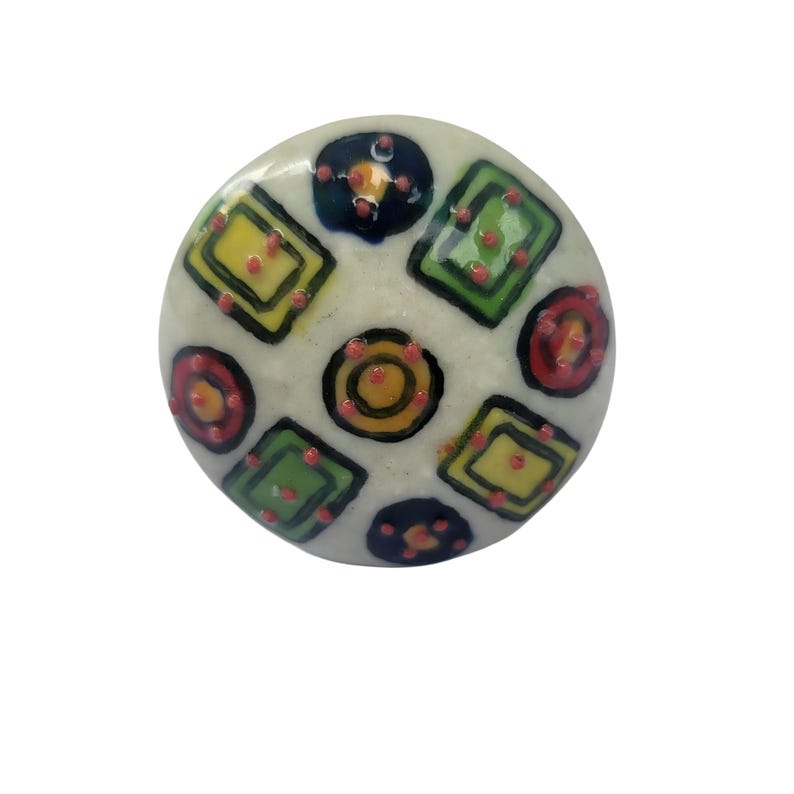 Peut inclure: Bouton de meuble rond en c&eacute;ramique blanc cass&eacute;, orn&eacute; d'un motif g&eacute;om&eacute;trique color&eacute;. Le design comprend des carr&eacute;s, des cercles et des points jaunes, verts, bleus et rouges, soulign&eacute;s de noir. Pour armoire ou tiroir.
