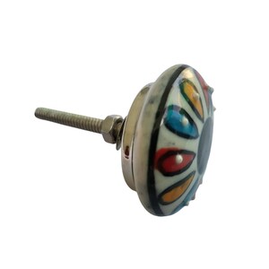 K&ouml;nnte beinhalten: Ein dekorativer Keramik-Schrankknopf mit einem farbenfrohen Blumendesign. Der Knopf hat eine cremefarbene Basis mit einem Muster aus roten, blauen, gelben und gr&uuml;nen Tropfenformen. Eine silberfarbene Metallschraube und -mutter sind angebracht.