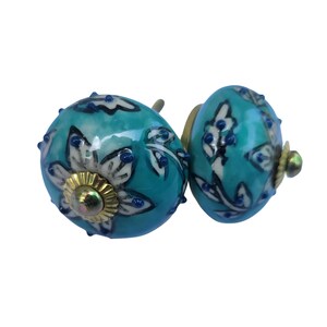 May include: Two turquoise ceramic cabinet knobs with floral designs. The knobs have a round shape, with white and black floral patterns and small blue accents. The base is gold-colored.