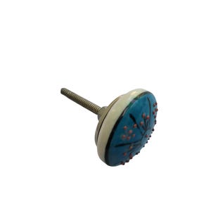 May include: A decorative cabinet knob with a round, turquoise face featuring a black floral design and small red accents. The knob has a cream-colored border and a metal screw for attachment.