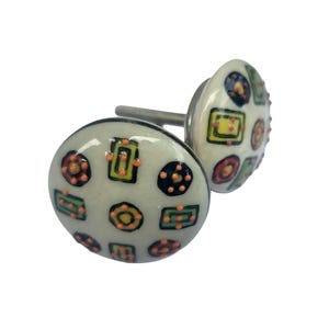 May include: Two decorative ceramic cabinet knobs. The knobs are round with a cream-colored background and feature a colorful geometric pattern of squares, circles, and rectangles in shades of green, yellow, orange, and navy blue.