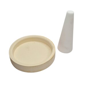 May include: A cream-colored ceramic bowl and a white conical object. The bowl is round with a shallow interior. The conical object is smooth and tapers to a rounded top. Both items are set against a white background.