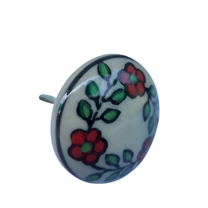 May include: A ceramic cabinet knob with a floral design. The knob is off-white with a black border and features red flowers and green leaves. The stem is a light green colour.