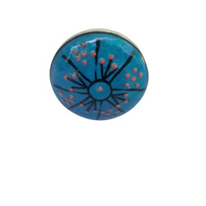 May include: A round, blue ceramic knob with a decorative design. The knob features a central black circle with radiating black lines and small red dots. The edges are a light cream color.