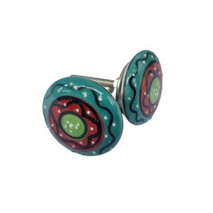 May include: Two decorative ceramic cabinet knobs. Each knob features a teal background with a red and green circular design. The design is accented with white dots and black wavy lines. The knobs are attached to a silver metal screw.
