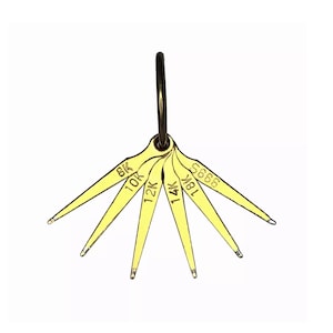 May include: A gold-colored jewelry gauge with a black ring at the top. The gauge has multiple fan-shaped blades, each labeled with a karat marking: 8K, 10K, 12K, 14K, 18K, and 9995. The background is plain white.