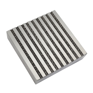 May include: A square, silver-colored metal block with a series of parallel, engraved lines. The lines alternate between solid silver and detailed, patterned black designs. The block is angled, showcasing its textured surface.