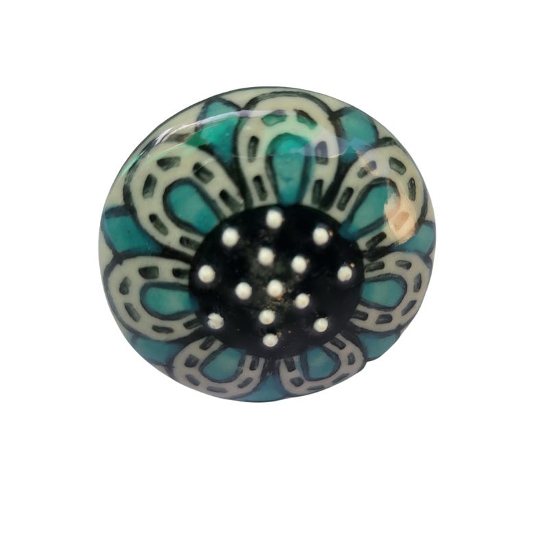 May include: A round ceramic knob with a floral design. The knob has a teal and white flower pattern with a black centre dotted with white spots. Likely for a cabinet or drawer. The knob is approximately 3 cm in diameter.