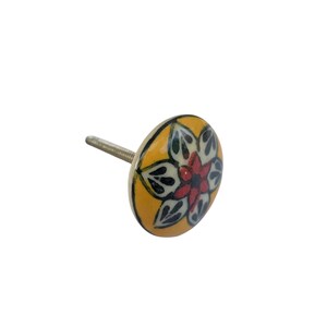 Op de afbeelding: Een decoratieve kastknop met een bloemmotief. De knop heeft een gele achtergrond met een rood, wit en zwart bloemmotief. De knop heeft een metalen schroef voor installatie.