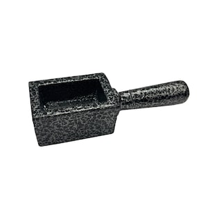 May include: A small, rectangular metal crucible with a textured, speckled finish. It has a short, rounded handle extending from the side. The crucible is dark gray with silver accents, and is designed for melting materials.