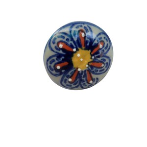 May include: A ceramic cabinet knob with a floral design. The knob features a light gray base with a blue, orange, and yellow flower pattern. The flower has a yellow center, orange petals, and blue outlines with white accents.