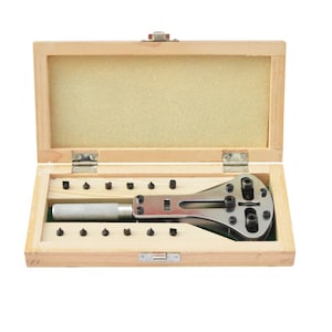 May include: A watch repair tool kit in an open wooden box. The kit includes a silver metal adjustable wrench, a knurled handle, and several small black pins. The box has a light brown exterior and a beige interior.
