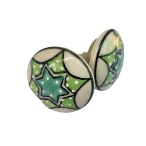 May include: Two ceramic cabinet knobs with a starburst design. The knobs are off-white with a green star and green and white accents. The star is outlined in black. The knobs are round and have a metal stem.