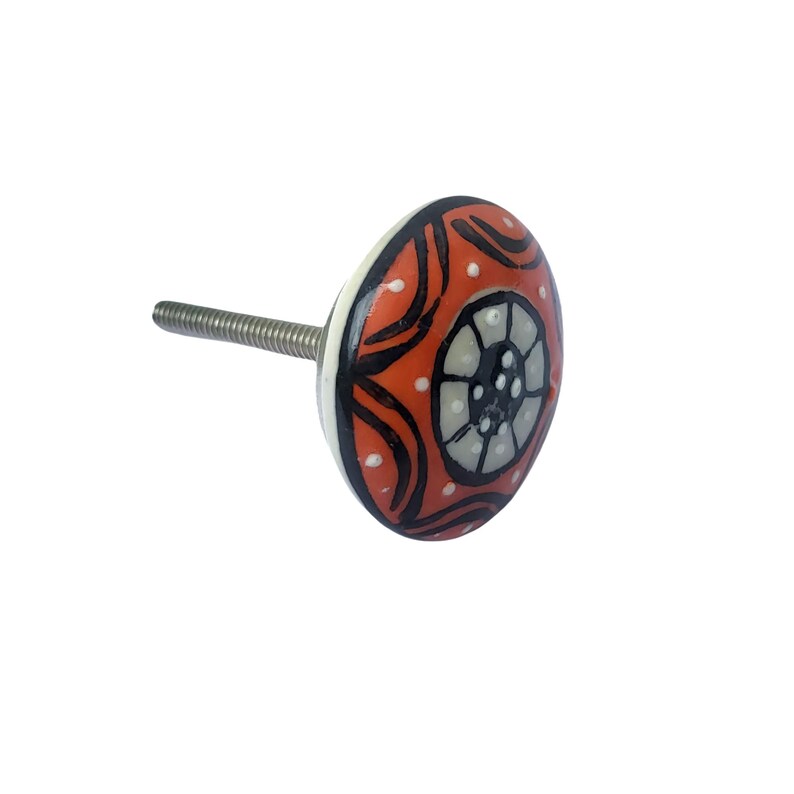 May include: A decorative ceramic cabinet knob with a vibrant orange background, black curved lines, white dots, and a central gray and white design. The knob has a silver threaded stem for easy installation. This hardware adds a pop of color.