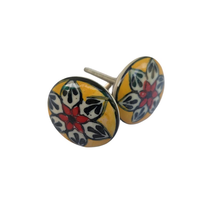Op de afbeelding: Twee decoratieve keramische kastknoppen. Elke knop heeft een gele achtergrond met een bloemenontwerp in rood, zwart en groen. De knoppen zijn ovaal van vorm en hebben een zilveren metalen schroef.