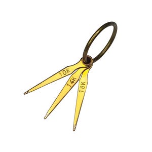 May include: A gold-toned metal tool with three prongs, each labeled with a different karat weight: 10K, 14K, and 18K. The tool is attached to a small ring.
