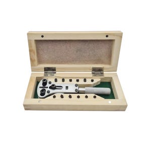 May include: A wooden box with a watch case opener inside. The opener has a metal handle and a variety of different sized holes for opening watch cases.