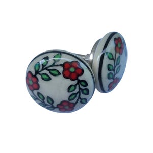May include: Two ceramic cabinet knobs with a floral design. The knobs are off-white with red flowers, green leaves, and black accents. The hardware is silver. These knobs would be a great addition to any cabinet or drawer.