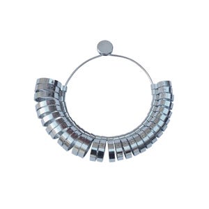 Op de afbeelding: Een zilveren ringmaat met een rond metalen frame en een reeks genummerde ringen. De ringen zijn in een cirkel gerangschikt, met de grootste ring bovenaan en de kleinste ring onderaan.
