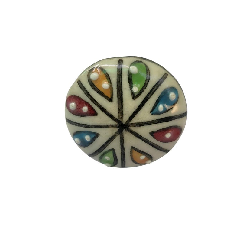 May include: A round, off-white ceramic knob with a colorful, geometric design. The design features segments in red, orange, green, and blue, separated by black lines. Each segment has a white dot. The knob is likely for a cabinet or drawer.