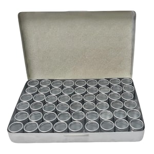 May include: A rectangular silver metal storage case with an open lid, containing a grid of fifty-six small, empty, cylindrical silver metal containers arranged in eight rows of seven, each with a circular opening and a metallic finish.