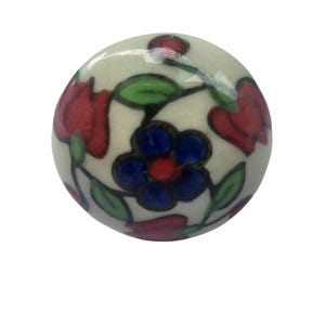 May include: A round, off-white ceramic cabinet knob with a floral design. The knob features red tulip-like flowers, blue and red centered flowers, and green leaves. The design is hand-painted with black outlines.