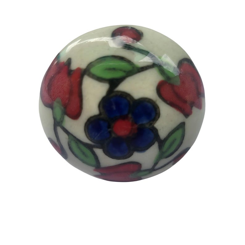 May include: A round, off-white ceramic cabinet knob with a floral design. The knob features red tulip-like flowers, blue and red centered flowers, and green leaves. The design is hand-painted with black outlines.